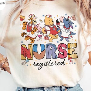 May include: Cream-colored t-shirt featuring a colorful graphic of Winnie the Pooh characters dressed as nurses. The word "NURSE" is displayed in large, multi-colored letters, with "registered" written below in script. The design includes medical symbols.