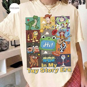 May include: A cream-colored t-shirt featuring a graphic print of various Toy Story characters, including Woody, Buzz Lightyear, and Rex. The design includes a green chalkboard with the text "Hi!" and the phrase "In My Tay Story Era" below.