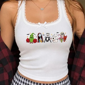 May include: A white ribbed tank top featuring a lace trim along the neckline and a small white bow at the center. The front displays a graphic print of various characters from The Nightmare Before Christmas, including Jack Skellington, Sally, and Zero.
