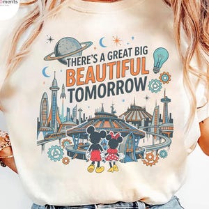 May include: A cream-colored t-shirt featuring a graphic print of a futuristic city with a monorail, a planet, a lightbulb, and gears. Two cartoon mice stand in the foreground. The text reads, There's a great big beautiful tomorrow.
