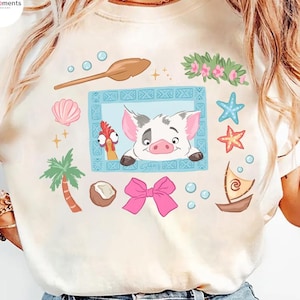 May include: Cream-colored t-shirt featuring a cartoon illustration of a pig and a rooster in a blue frame, surrounded by tropical elements like a palm tree, seashells, and a pink bow. The shirt has the text "HisMoments" on the upper left.