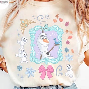 May include: Cream-colored t-shirt featuring a colorful graphic of Olaf from Frozen. Olaf is in a decorative frame, holding a blue bird. The design includes snowflakes, a pink bow, and other winter-themed elements. The shirt has a slightly faded, vintage look.