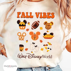 May include: Cream-colored t-shirt with the text "FALL VIBES" in orange. The shirt features various fall-themed graphics, including Mickey Mouse-shaped pumpkins, a football, and autumn leaves. The words "Walt Disney World" are also printed on the shirt.