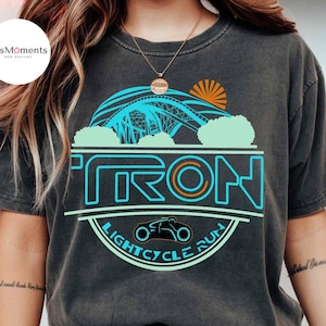 May include: Dark gray t-shirt featuring a graphic design with the word "TRON" in teal and a light cycle illustration. The design includes a bridge, sun, and the words "LIGHTCYCLE RUN". The shirt has a vintage, worn look.