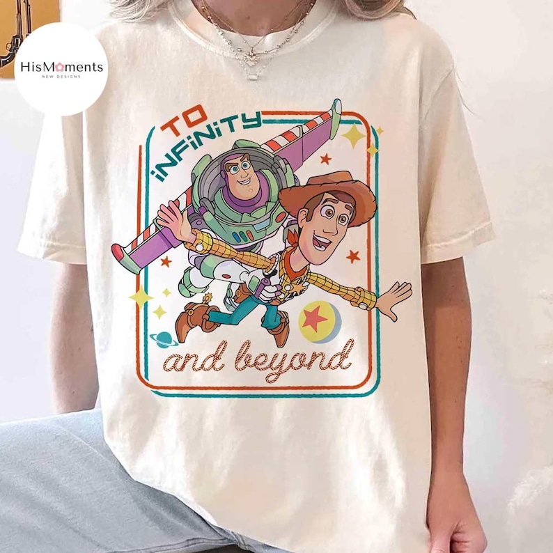 May include: Cream-colored t-shirt featuring a graphic of Buzz Lightyear and Woody from Toy Story, with the text "To Infinity and Beyond" in a retro design. The characters are depicted in mid-air, with a colorful border.