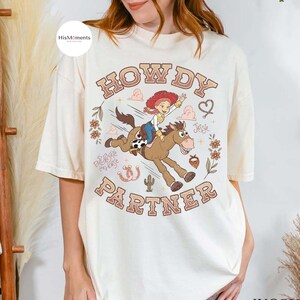 May include: An off-white t-shirt featuring a graphic of a cowgirl riding a brown horse. The text "HOWDY PARTNER" is printed in brown letters above and below the illustration. Additional details include a cactus, flowers, and a horseshoe.