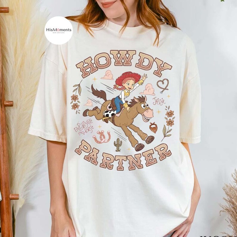 May include: An off-white t-shirt featuring a graphic of a cowgirl riding a brown horse. The text "HOWDY PARTNER" is printed in brown letters above and below the illustration. Additional details include a cactus, flowers, and a horseshoe.