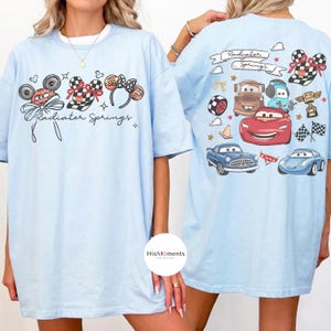Może przedstawiać: Jasnoniebieskie koszulki z grafiką z przodu i z tyłu. Z przodu znajdują się uszy Myszki Miki, kraciasta kokarda i napis "Radiator Springs". Z tyłu znajdują się kresk&oacute;wkowe samochody i napis "Radiator Springs."