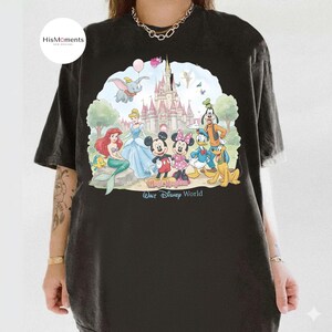 Pu&ograve; includere: Una t-shirt nera con una grafica colorata di vari personaggi dei cartoni animati davanti a un castello. La stampa include Topolino, Minnie, Paperino, Pippo, Pluto, Ariel, Cenerentola e Dumbo, con il testo Magic Kingdom Walt Disney World.