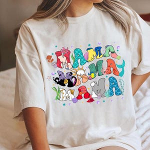 May include: White t-shirt with a colorful graphic of the word "Mama" made up of characters from the Little Mermaid movie. The characters include Ariel, Ursula, Prince Eric, and Sebastian.