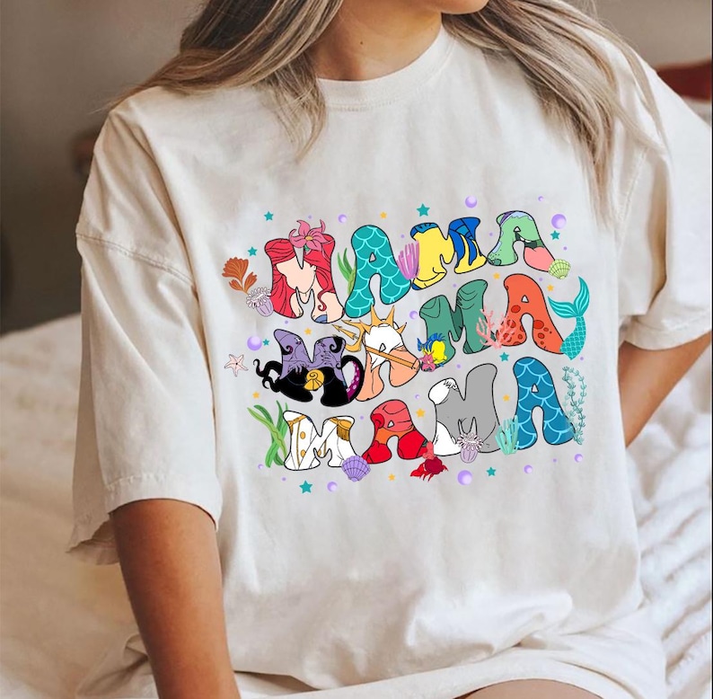 May include: White t-shirt with a colorful graphic of the word "Mama" made up of characters from the Little Mermaid movie. The characters include Ariel, Ursula, Prince Eric, and Sebastian.