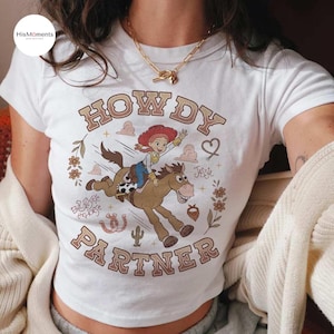 May include: A white short-sleeved t-shirt featuring a graphic of Jessie riding Bullseye the horse. The text reads "HOWDY PARTNER" in brown letters, with "Jessie" and "Bullseye the horse" written in script. Floral accents, a horseshoe, and a cactus are included.