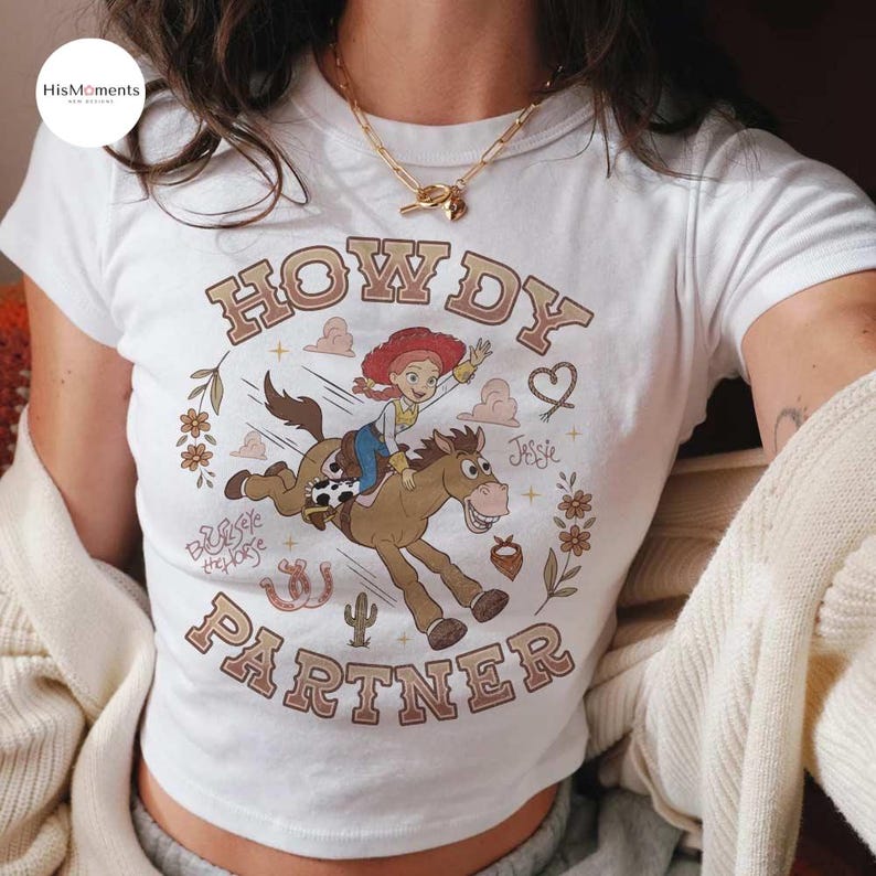 May include: A white short-sleeved t-shirt featuring a graphic of Jessie riding Bullseye the horse. The text reads "HOWDY PARTNER" in brown letters, with "Jessie" and "Bullseye the horse" written in script. Floral accents, a horseshoe, and a cactus are included.