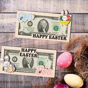 Könnte beinhalten: Zwei hölzerne Geldhalter mit einem Design aus 1-Dollar- und 2-Dollar-Scheinen. Die Scheine zeigen den Text "Frohe Ostern" und den Namen "Eva". Die Halter sind mit Osternhase- und Eiergrafiken verziert.
