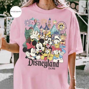 May include: A light pink t-shirt featuring a colorful graphic of Mickey Mouse, Minnie Mouse, Donald Duck, Daisy Duck, Goofy, Pluto, and Chip 'n Dale in front of a castle and ferris wheel. The text Disneyland Est. 1955 is printed below the characters.