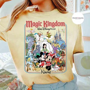 Puede incluir: Camiseta amarilla clara con un dise&ntilde;o gr&aacute;fico colorido de personajes de Disney, incluyendo Mickey y Minnie Mouse, con el texto "Magic Kingdom Walt Disney World" y un espacio para un nombre. El dise&ntilde;o est&aacute; enmarcado con un borde rojo y azul.