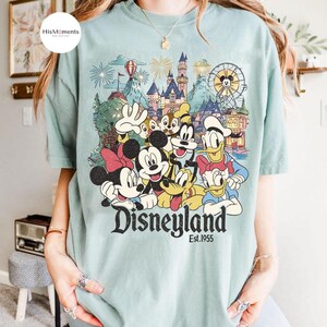 May include: A light green t-shirt featuring a graphic print of Mickey Mouse, Minnie Mouse, Donald Duck, Daisy Duck, Goofy, Pluto, and Chip 'n Dale in front of a castle and Ferris wheel. The text Disneyland Est. 1955 is printed below the characters.