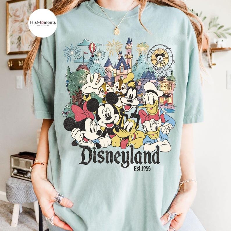 May include: A light green t-shirt featuring a graphic print of Mickey Mouse, Minnie Mouse, Donald Duck, Daisy Duck, Goofy, Pluto, and Chip 'n Dale in front of a castle and Ferris wheel. The text Disneyland Est. 1955 is printed below the characters.