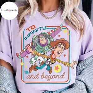 May include: A light purple t-shirt featuring a graphic of Buzz Lightyear and Woody from Toy Story, with the text "TO INFINITY and beyond" in a retro style. The design includes stars and a planet.