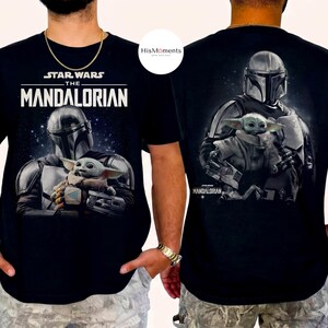 May include: Black t-shirt featuring a graphic of The Mandalorian and Grogu from Star Wars. The front of the shirt displays the text "STAR WARS THE MANDALORIAN" above the image. The back of the shirt also features the Mandalorian and Grogu.