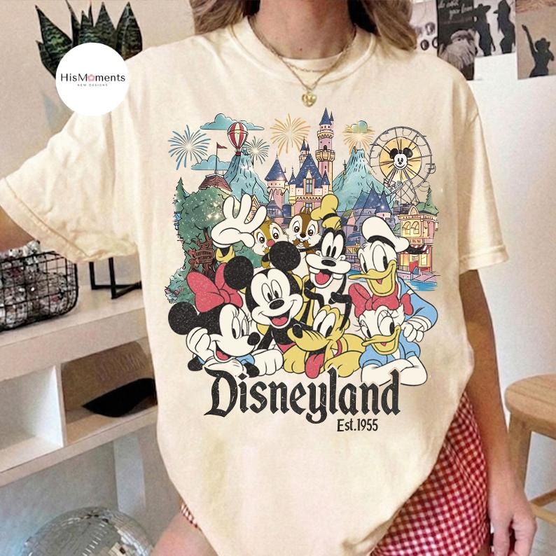 May include: A cream-colored short-sleeve t-shirt featuring a colorful graphic print of Mickey Mouse, Minnie Mouse, Donald Duck, Daisy Duck, Goofy, Pluto, and Chip 'n Dale in front of a castle, with the text Disneyland Est. 1955 printed below.