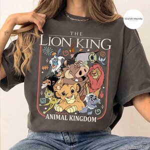 May include: Dark gray t-shirt featuring a colorful graphic of characters from The Lion King animated film. The design includes Simba, Timon, Pumbaa, and other characters, with the text "THE LION KING ANIMAL KINGDOM".