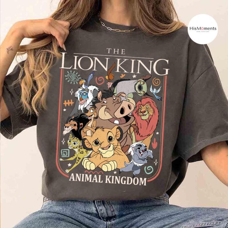 May include: Dark gray t-shirt featuring a colorful graphic of characters from The Lion King animated film. The design includes Simba, Timon, Pumbaa, and other characters, with the text "THE LION KING ANIMAL KINGDOM".