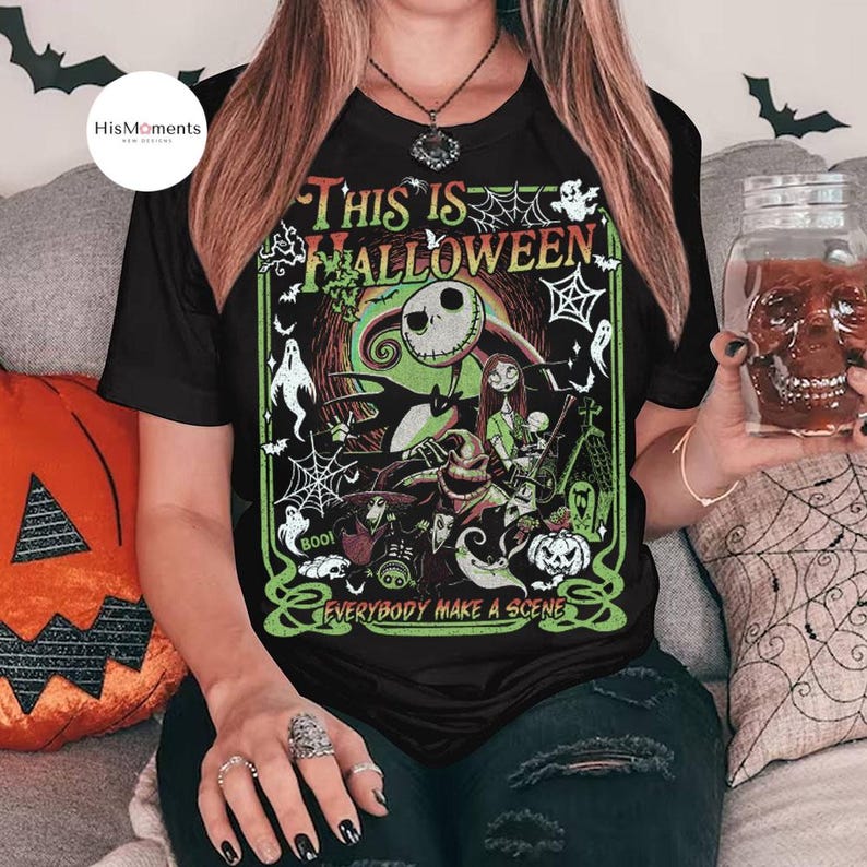 K&ouml;nnte beinhalten: Schwarzes T-Shirt mit einem Halloween-Motiv mit Jack Skellington und Sally. Das Design enth&auml;lt den Text "THIS IS HALLOWEEN" und "EVERYBODY MAKE A SCENE" in Gr&uuml;n und Wei&szlig;. Ein dekoratives, sch&auml;delartiges Glas wird in der Hand gehalten.