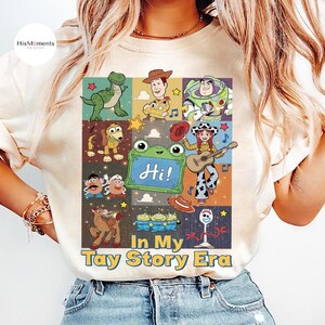 May include: A cream-colored t-shirt featuring a grid of cartoon characters from the Toy Story franchise, including a green dinosaur, a cowboy, a space ranger, a dog, a potato toy, a frog, a cowgirl with a guitar, and a spork. Text reads In My Tay Story Era.