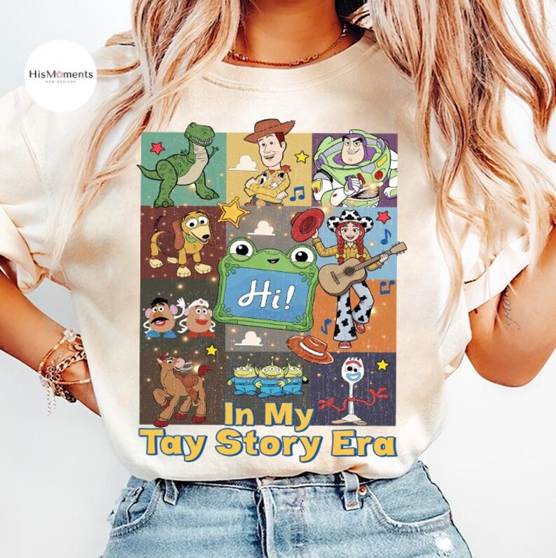 May include: A cream-colored t-shirt featuring a grid of cartoon characters from the Toy Story franchise, including a green dinosaur, a cowboy, a space ranger, a dog, a potato toy, a frog, a cowgirl with a guitar, and a spork. Text reads In My Tay Story Era.