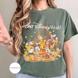 Op de afbeelding: Groen t-shirt met de tekst "Walt Disney World" en een afbeelding van Disney-personages, waaronder Mickey en Minnie Mouse, Donald Duck en Goofy, met pompoenen en herfstbladeren.