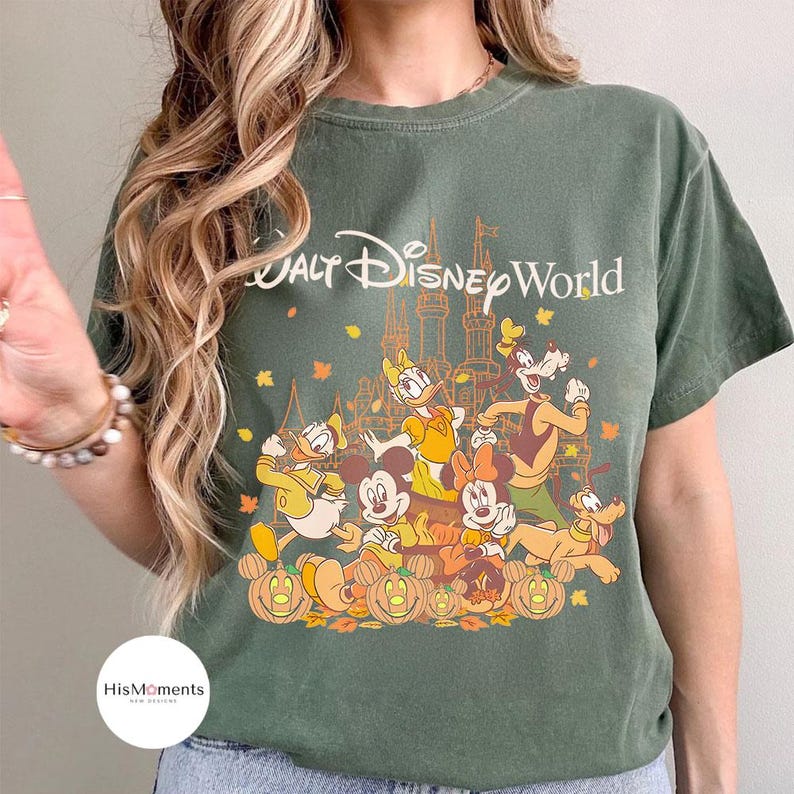 Op de afbeelding: Groen t-shirt met de tekst "Walt Disney World" en een afbeelding van Disney-personages, waaronder Mickey en Minnie Mouse, Donald Duck en Goofy, met pompoenen en herfstbladeren.