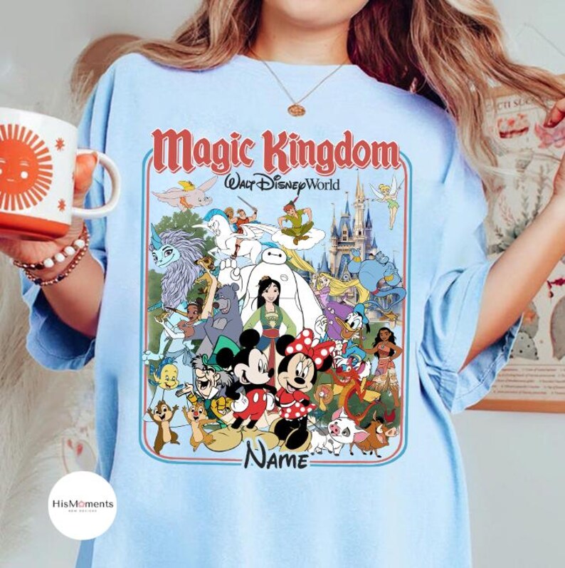 Puede incluir: Camiseta azul claro con un dise&ntilde;o de personajes de Disney y el texto "Magic Kingdom Walt Disney World". El dise&ntilde;o incluye a Mickey y Minnie Mouse, y otros personajes. La palabra "Name" tambi&eacute;n es visible.