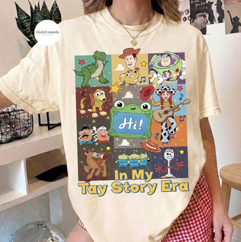 May include: A cream-colored t-shirt featuring a graphic print of various Toy Story characters, including Woody, Buzz Lightyear, and Rex. The design includes a green chalkboard with the text "Hi!" and the phrase "In My Tay Story Era" below.