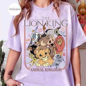 May include: A light purple t-shirt featuring a colorful graphic of characters from The Lion King. The design includes the text "THE LION KING" above the words "ANIMAL KINGDOM". The shirt has short sleeves and a crew neck.