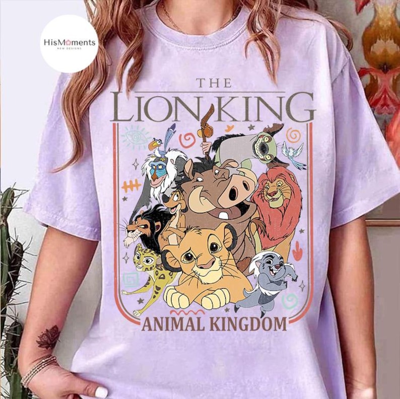 May include: A light purple t-shirt featuring a colorful graphic of characters from The Lion King. The design includes the text "THE LION KING" above the words "ANIMAL KINGDOM". The shirt has short sleeves and a crew neck.
