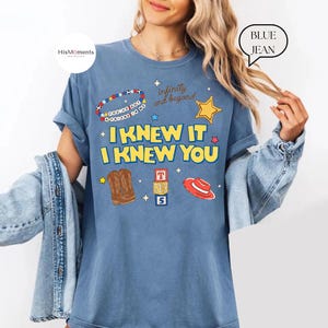 K&ouml;nnte beinhalten: Ein blaues T-Shirt in Jeansoptik mit dem Text I KNEW IT I KNEW YOU in gelben Buchstaben. Die Grafik zeigt ein Perlenarmband, einen gelben Stern, braune Cowboystiefel, einen Stapel Baukl&ouml;tze mit den Buchstaben T S 5 und einen roten Cowboyhut.