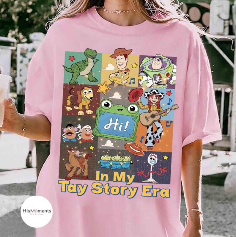 May include: A light pink t-shirt featuring a grid of Toy Story characters including Rex, Woody, Buzz Lightyear, Slinky Dog, Mr. and Mrs. Potato Head, Bullseye, Jessie, the Aliens, and Forky, with the text "Hi!" and "In My Tay Story Era."