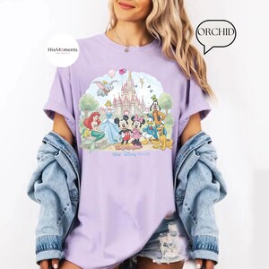 Pu&ograve; includere: Una t-shirt color viola chiaro con una stampa grafica di un castello e vari personaggi dei cartoni animati tra cui Topolino, Minnie, Paperino, Pippo, Pluto, Ariel e Dumbo. Il testo "Magic Kingdom" e "Walt Disney World" &egrave; stampato sotto.