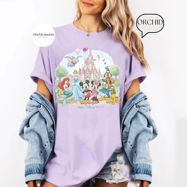 Pu&ograve; includere: Una t-shirt color viola chiaro con una stampa grafica di un castello e vari personaggi dei cartoni animati tra cui Topolino, Minnie, Paperino, Pippo, Pluto, Ariel e Dumbo. Il testo "Magic Kingdom" e "Walt Disney World" &egrave; stampato sotto.