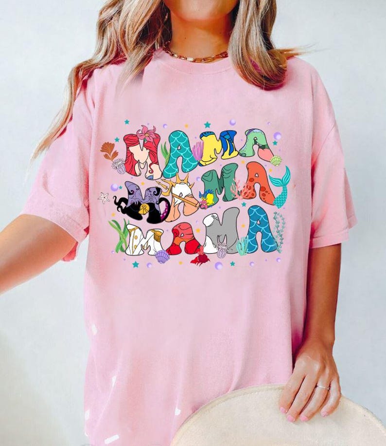 May include: Pink t-shirt with a graphic of the word "Mama" in colorful letters with various Disney characters from The Little Mermaid.