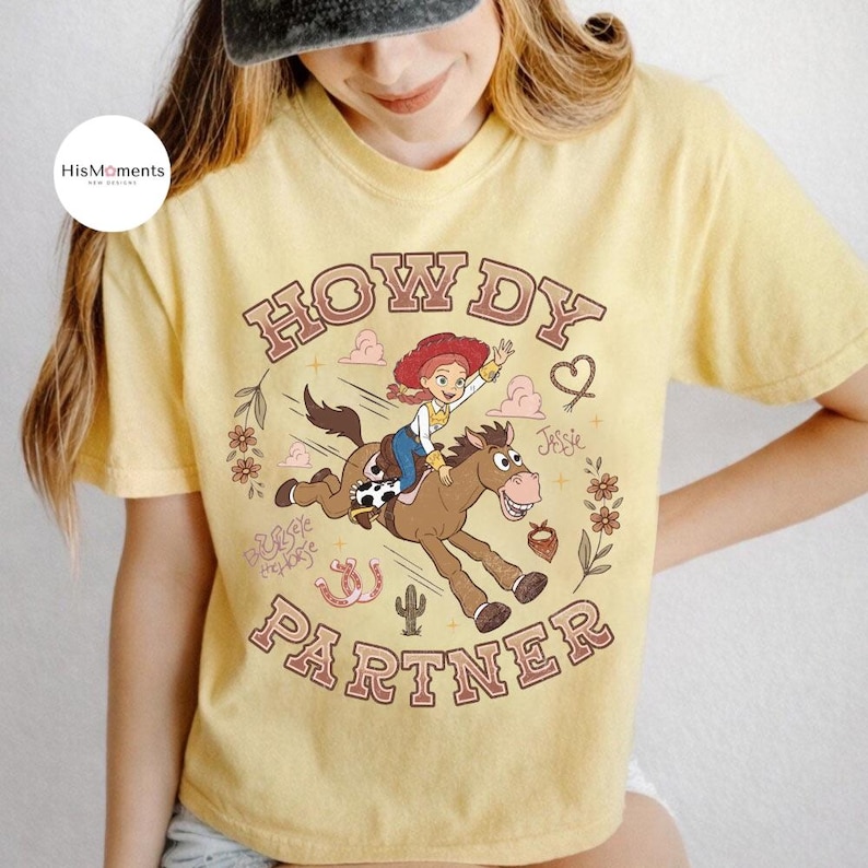 May include: A pale yellow t-shirt featuring a graphic of Jessie riding Bullseye from Toy Story. The text reads HOWDY PARTNER, with decorative elements including a cactus, flowers, a horseshoe, and a heart. The design has a faded, vintage aesthetic.