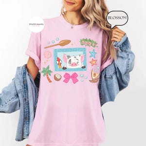 May include: A light pink t-shirt featuring a cartoon illustration of a pig and a rooster within a blue frame, surrounded by tropical elements like a palm tree, seashells, and stars. The shirt has a relaxed fit.
