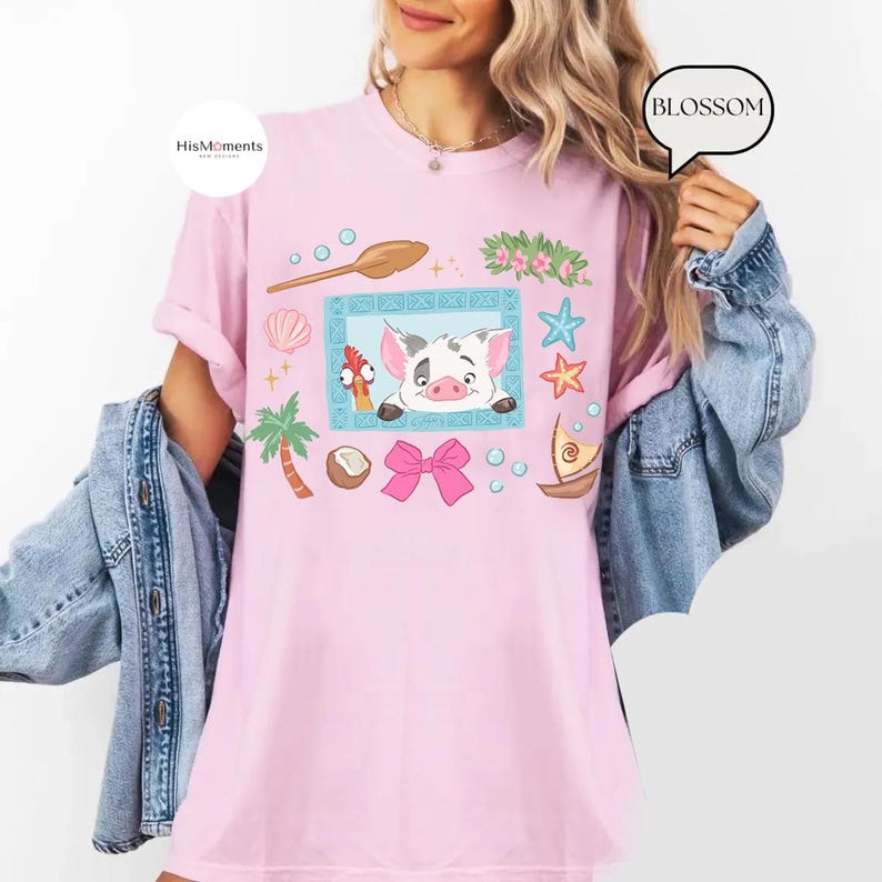 May include: A light pink t-shirt featuring a cartoon illustration of a pig and a rooster within a blue frame, surrounded by tropical elements like a palm tree, seashells, and stars. The shirt has a relaxed fit.