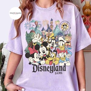 May include: A light purple t-shirt featuring a graphic print of Mickey Mouse, Minnie Mouse, Donald Duck, Daisy Duck, Goofy, Pluto, and Chip 'n Dale in front of a castle, a Ferris wheel, and fireworks with the text Disneyland Est. 1955.