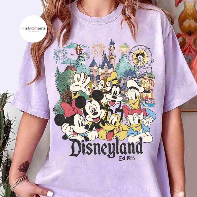 May include: A light purple t-shirt featuring a graphic print of Mickey Mouse, Minnie Mouse, Donald Duck, Daisy Duck, Goofy, Pluto, and Chip 'n Dale in front of a castle, a Ferris wheel, and fireworks with the text Disneyland Est. 1955.