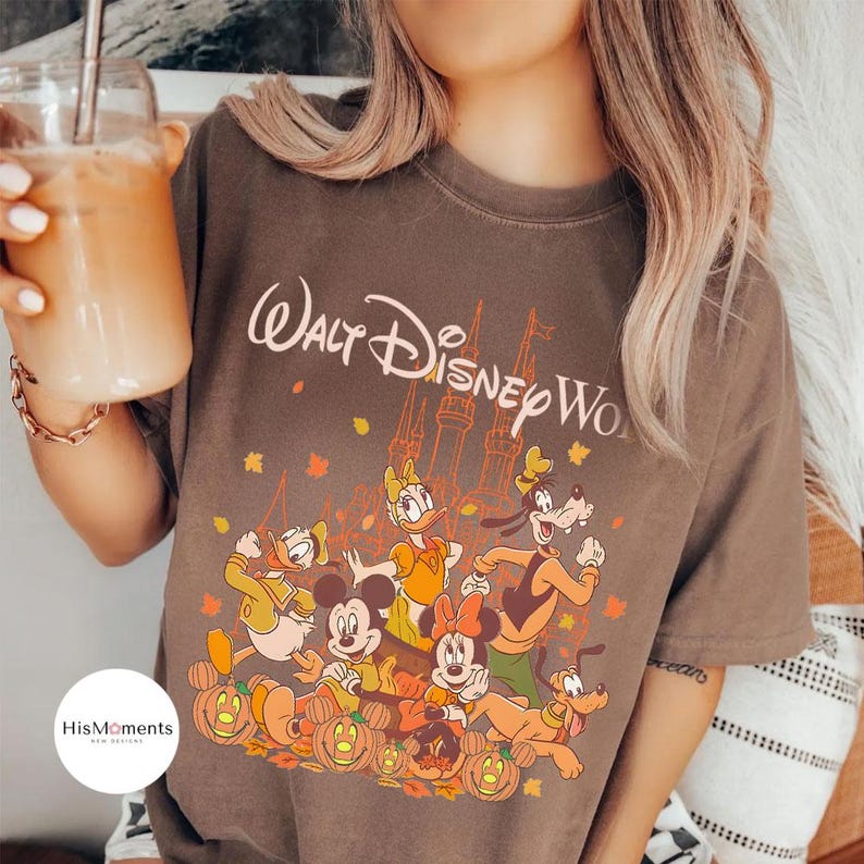 Op de afbeelding: Bruin T-shirt met de tekst "Walt Disney World" en een kasteelafbeelding. Het ontwerp toont Disney-personages zoals Mickey en Minnie Mouse, Donald Duck en Goofy, omringd door pompoenen en herfstbladeren.