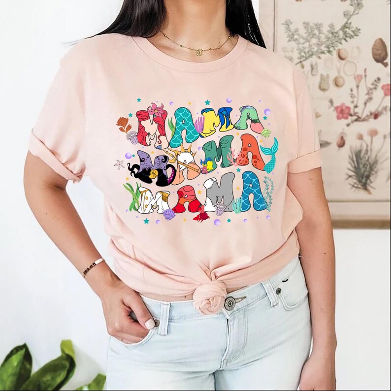 May include: A pink t-shirt with a graphic of the word "mama" spelled out with various Disney characters from The Little Mermaid. The characters are in different colors and are surrounded by stars and seashells.