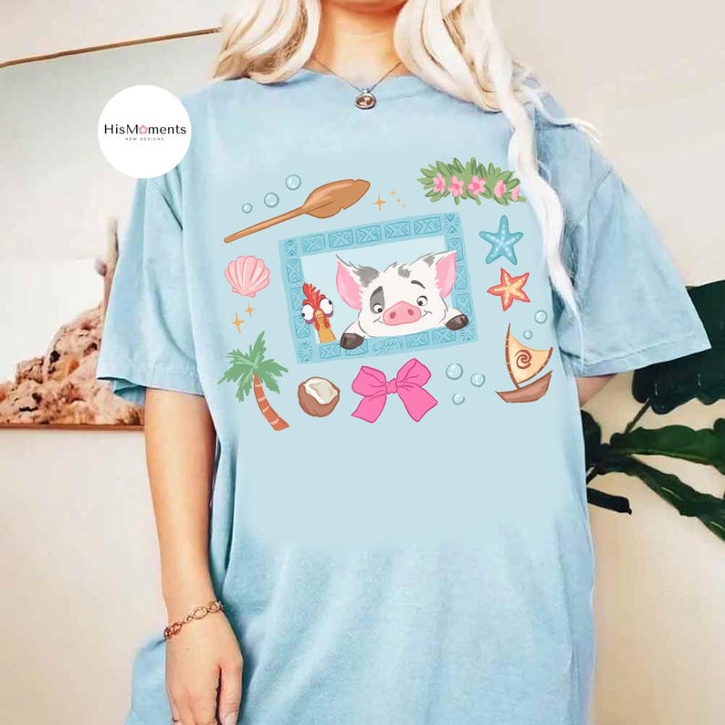 May include: Light blue t-shirt featuring a cartoon graphic with a pig, chicken, and other tropical elements. The design includes a wooden spoon, seashells, and a pink bow. The shirt has the text "HisMoments" on the upper left.