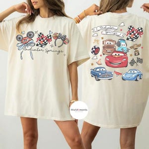 Może przedstawiać: Kremowa koszulka z nadrukiem z przodu przedstawiającym uszy Myszki Miki, koła samochod&oacute;w wyścigowych i napis "Radiator Springs". Tył koszulki przedstawia r&oacute;żne kresk&oacute;wkowe samochody i napis "Radiator Springs".